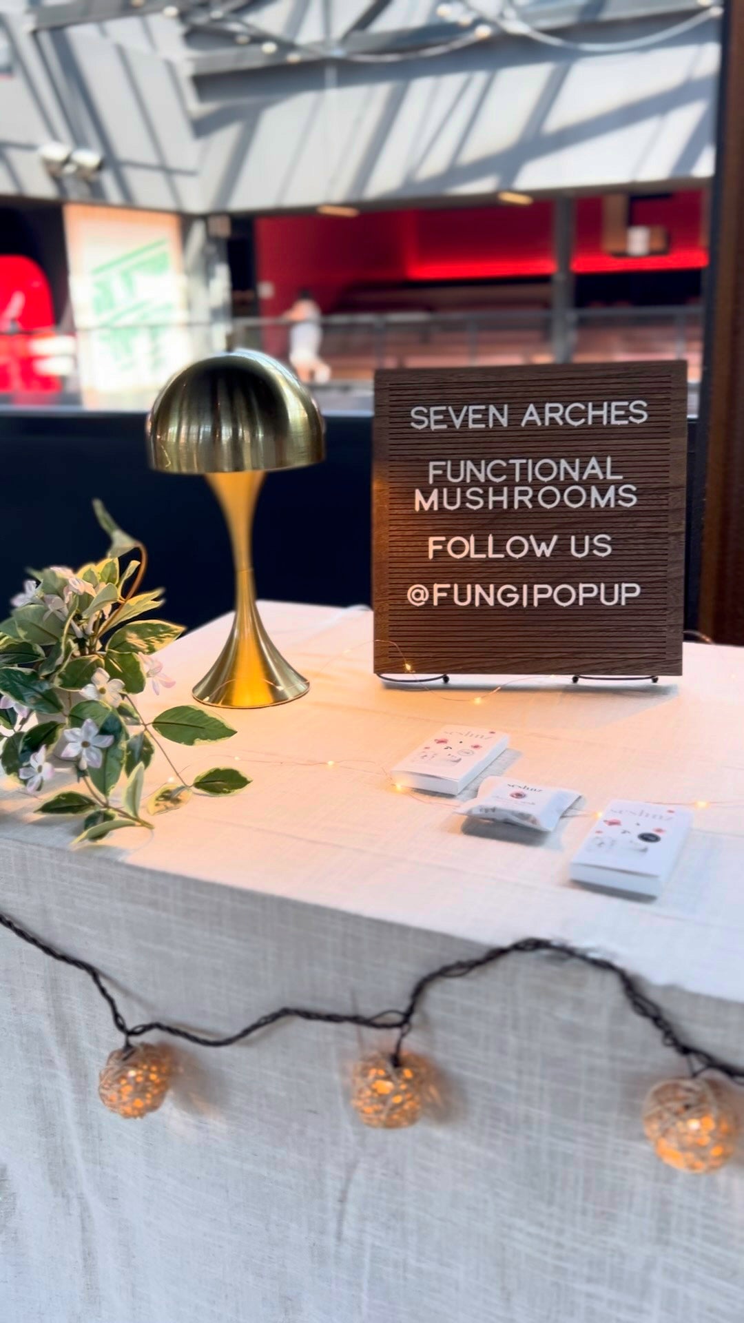 Real FungiPopUp event display with Seven Arches functional mushrooms sign, @FUNGIPOPUP lettering, tabletop products, gold lamp, and string lights